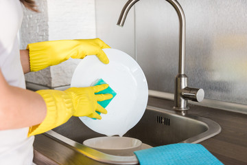Fototapeta na wymiar A young woman in yellow gloves washes dishes with a sponge in the sink. House professional cleaning service.