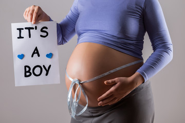 Image of close up stomach of pregnant woman with blue ribbon holding paper with text it's a boy on gray background.