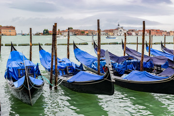 Empty gondolas docked between wooden mooring poles covered in tarpaulin in rainy November season. Dome top building in background is Le Zitelle Church on Giudecca another island in the Venetian lagoon