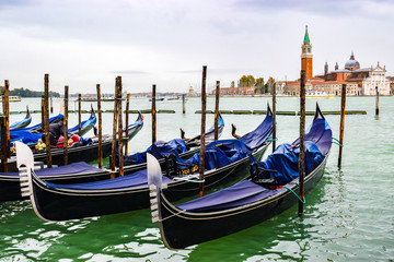 Fototapeta na wymiar Covered gondolas docked on water between wooden mooring poles. Bell tower in background is of 16th century Palladian architecture Church of San Giorgio Maggiore on San Giorgio Maggiore island, Venice.