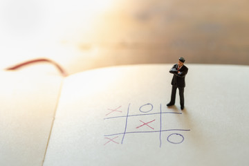 Business direction and planning concept. Businessman miniature figure standing and thinking on paper with OX (tic tac toe) board game.