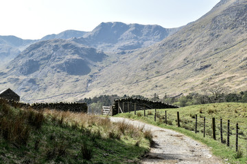Fototapeta na wymiar Gravel path leading up to stone wall with wooden gate. Stunning nature in the background. Slightly toned down colors. Grisedale pike, Lake District, England, UK -Image