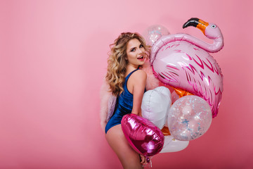 Cheerful curly girl in dark-blue velvet combination poses with a toy inflatable flamingo on pink background. Portrait of gorgeous blonde young woman holding colorful party balloons and having fun - obrazy, fototapety, plakaty
