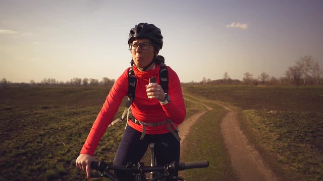 Athletic Caucasian woman eats protein bar ride on mountain bike on nature. Young sporty woman athlete in helmet resting while biting nutritional bar. Fitness woman eating energy snack outdoor