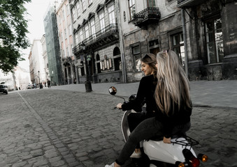 Fototapeta premium Young and beautiful girls in sunglasses riding a scooter. Portrait of pretty ladies riding a motorbike on old downtown street. Summer vacation. Girls in a black t-shirts on a vintage scooter