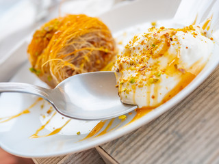 Close up of a succulent Greek dessert. Greek kataifi with honey yogurt and grated pistachios on a white plate and a blurred background. Horizontal view