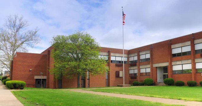 A daytime Spring establishing shot of a typical two-story red brick school building in a small midwestern town. Partially overcast. American flag waving on flagpole in front. Winter/Summer matching.