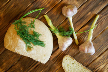 Garlic, dill sprigs and a white round bread bun are lying on a wooden table made of pine boards. Summer still life