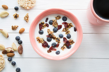 A bowl with yogurt, cornflakes, blueberry and nuts on white rustic table. A cup of black coffee. 