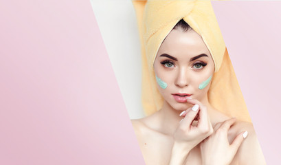 Portrait of beautiful woman with healthy glow perfect smooth skin. Model with towel on head look into the hole of colored pink paper. Young girl enjoys cream facial mask.