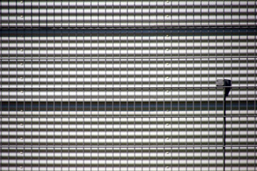 Das Detail eines Gitterzaunes vor der Wellblechfassade eines &ouml;ffentlichen Geb&auml;udes. ..