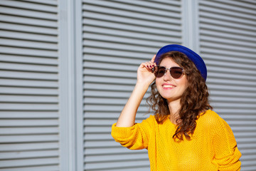 Street portrait of attractive woman wearing blue hat and yellow sweater posing in sun rays. Empty space