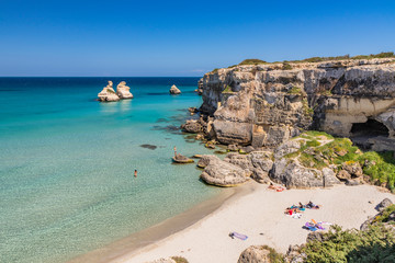 The bay of Torre dell'Orso, with its high cliffs, in Salento, Puglia, Italy. Turquoise sea and blue sky. A beach of fine white and pink sand. The stacks called the Two Sisters. Tourists sunbathe.