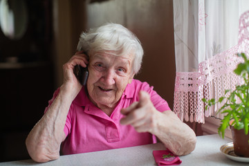 An old woman talking on the phone sitting at a table in his house.