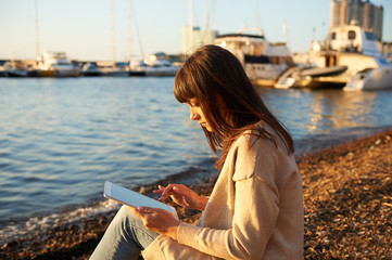 young woman sitting on the beach, at sunset, reading from the tablet