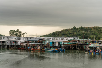 Fototapeta na wymiar Hong Kong, China - March 7, 2019: White Fishermen houses on stilts on Tai O River with green hill in back under gray sky with yellow horizon. Color added by sloops, buoys and tarps.