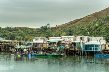 Fototapeta na wymiar Hong Kong, China - March 7, 2019: White Fishermen houses on stilts on Tai O River with green hill in back under gray sky. Color added by sloops, buoys and tarps.