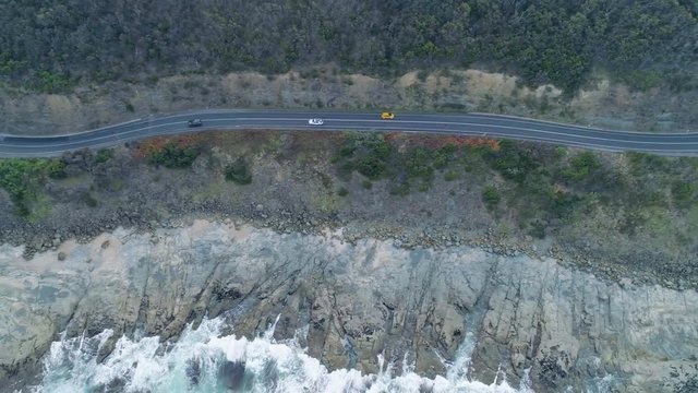 Tracking aerial shot of cars driving on narrow winding road on rugged ocean coastline