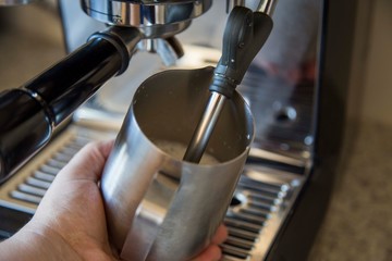 Close up of someone steaming and frothing the milk for a flat white at home using a brushed steel jug and a black espresso machine on a grey kitchen bench.