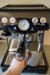 Steaming and frothing the milk for a flat white using a brushed steel jug and a black espresso machine on a grey kitchen bench.
