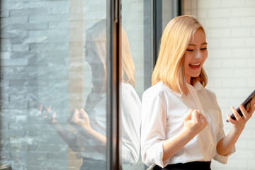 Fototapeta premium Young businesswoman standing near the office window smiling arms up celebrating with mobile phone success work at the office, business concept, success concept