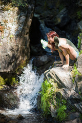 Red-haired woman in shorts doing yoga at the waterfall. The girl is standing in the pose of a firefly, titibhasana