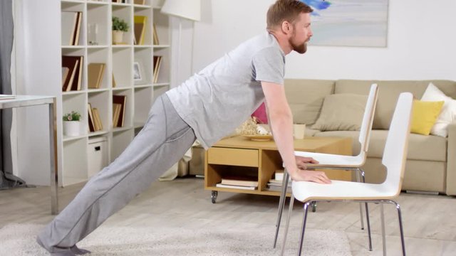 Strong man in sportswear using two chairs for doing push-ups while training in the living room at home