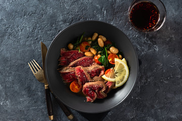 Dinner plate with beef steak with white beans, spinach and tomatoes on dark stone background top view with glass of red wine