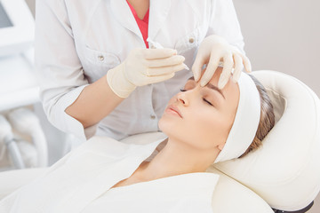 Unidentified master cosmetologist makes botox injections into the eyebrow of a beautiful young woman client. The concept of rejuvenating aesthetic procedures - obrazy, fototapety, plakaty