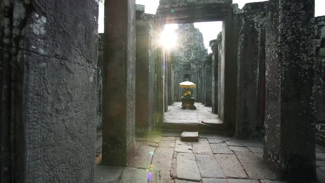 Long corridor in ancient Bayon temple with carvings on walls, Buddha statue in gold clothes and sun rising above it. Angkor Wat complex. Cambodia