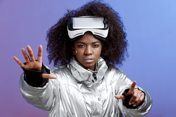 Fototapeta na wymiar Modern curly brown-haired girl dressed in a silver-colored jacket wearing on her head the virtual reality glasses poses in the studio on neon background