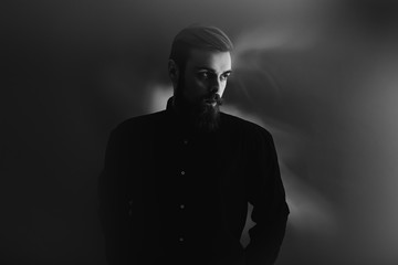 Black and white photo of a stylish man with a beard dressed in the black shirt stands in the fog