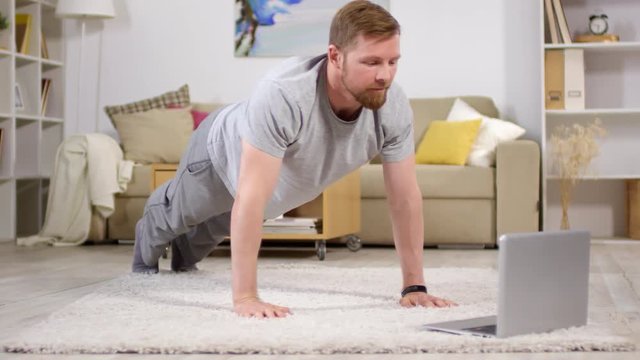 Caucasian man in sportswear doing push-ups on the floor in living room and watching workout video on laptop