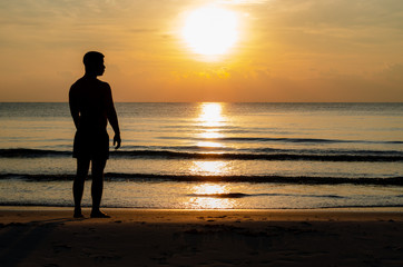 The silhouette photo of a man standing alone on the beach enjoy sunrise moment with the reflection on the sea in summer season.