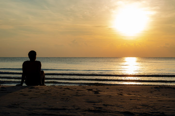 The silhouette photo of a man sitting alone on the beach enjoy sunrise moment with the reflection on the sea in summer season.