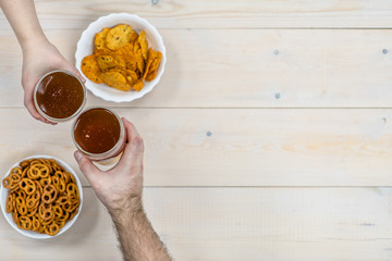 Hands holding glass mug of beer with pretzels and chips on light wooden background. Empty space for text. Top view