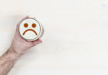 man's hand holds a mug of beer with sad face on the foam. Light wooden background. Empty space for text