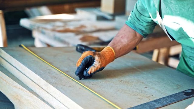 Worker in special uniform measures the stone slabs in factory. Man with the tape measure ticks off by marker on a slab in manufacture.