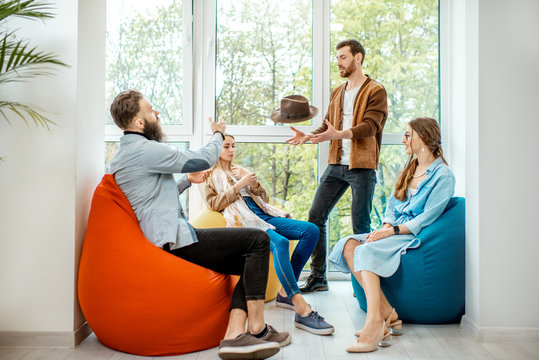 Group of diverse people having a conversation during the coffee break sitting on the colorful poufs near the window in the office