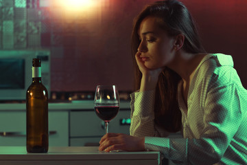 Lonely, sad, beautiful young woman with red wine is drinking alone in the kitchen at home in evening. Female alcoholism and alcohol addiction.