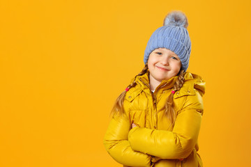 Closeup portrait of a cheerful little girl in jacket and knitted hat over yellow background. The child folded her arms and looks into the camera.