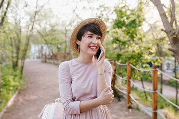Amazing girl with short dark hair speaking on phone and looking up with smile. Outdoor portrait of inspired young lady wearing summer hat and cute gown walking down the alley.