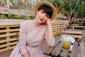 Obraz premium Lovely girl with nude makeup sitting at the wooden table propping face with hand. Portrait of charming young lady in straw hat and trendy pastel dress resting in outdoor cafe with glass of juice.