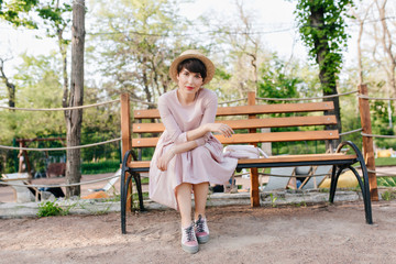 Good-looking young woman in interesting vintage outfit with white backpack posing, during rest in the park. Portrait of graceful girl with short haircut chilling on bench in warm spring day.