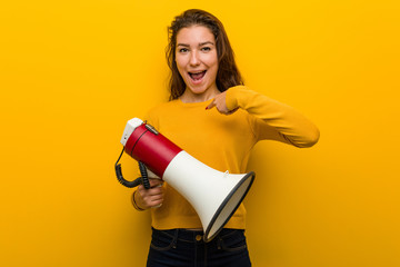 Young european woman holding a megaphone surprised pointing at herself, smiling broadly.