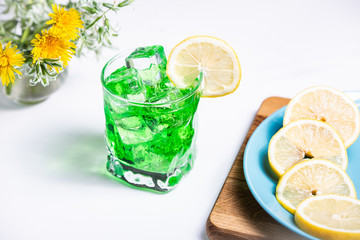 Transparent green drink in a glass with a slice of lemon and ice cubes. Lemon slices on a blue plate.