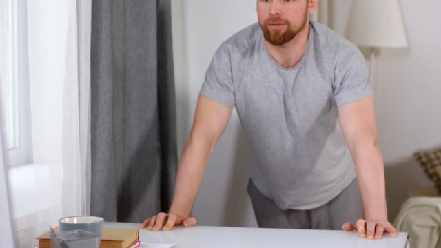 Tilt down shot of young bearded man doing table push-ups at home and watching movie on computer