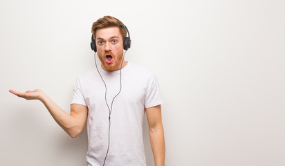 Young redhead man holding something on palm hand. Listening to music with headphones.