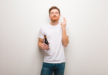 Young redhead man crossing fingers for having luck. Holding a beer.