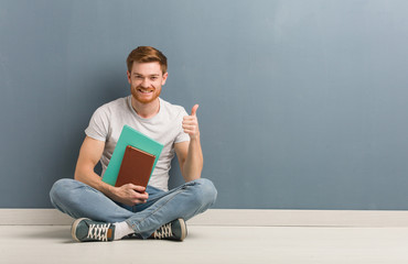 Young redhead student man sitting on the floor smiling and raising thumb up. He is holding books.